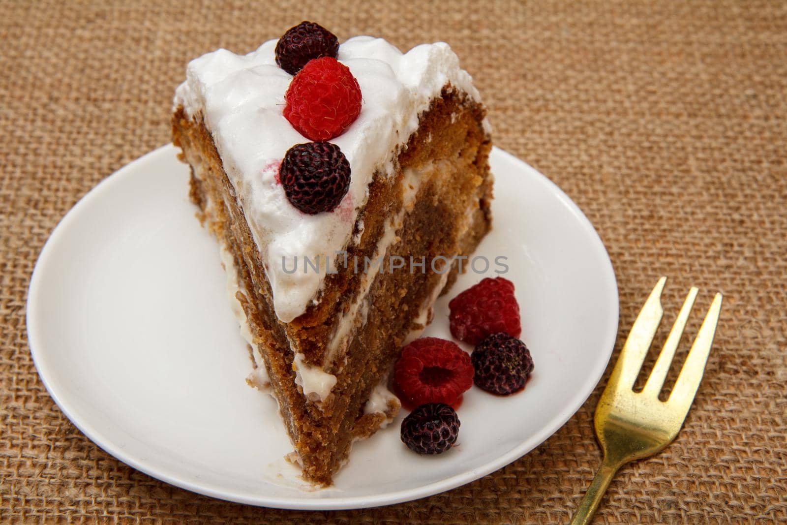 Slice of homemade biscuit cake decorated with whipped cream and raspberries on table with sackcloth