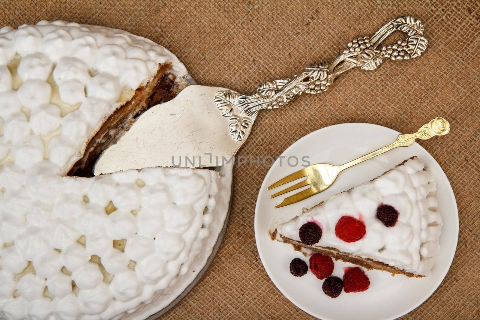 Sliced biscuit cake decorated with whipped cream and raspberries, silver cake lifter beside it on table with sackcloth. Top view