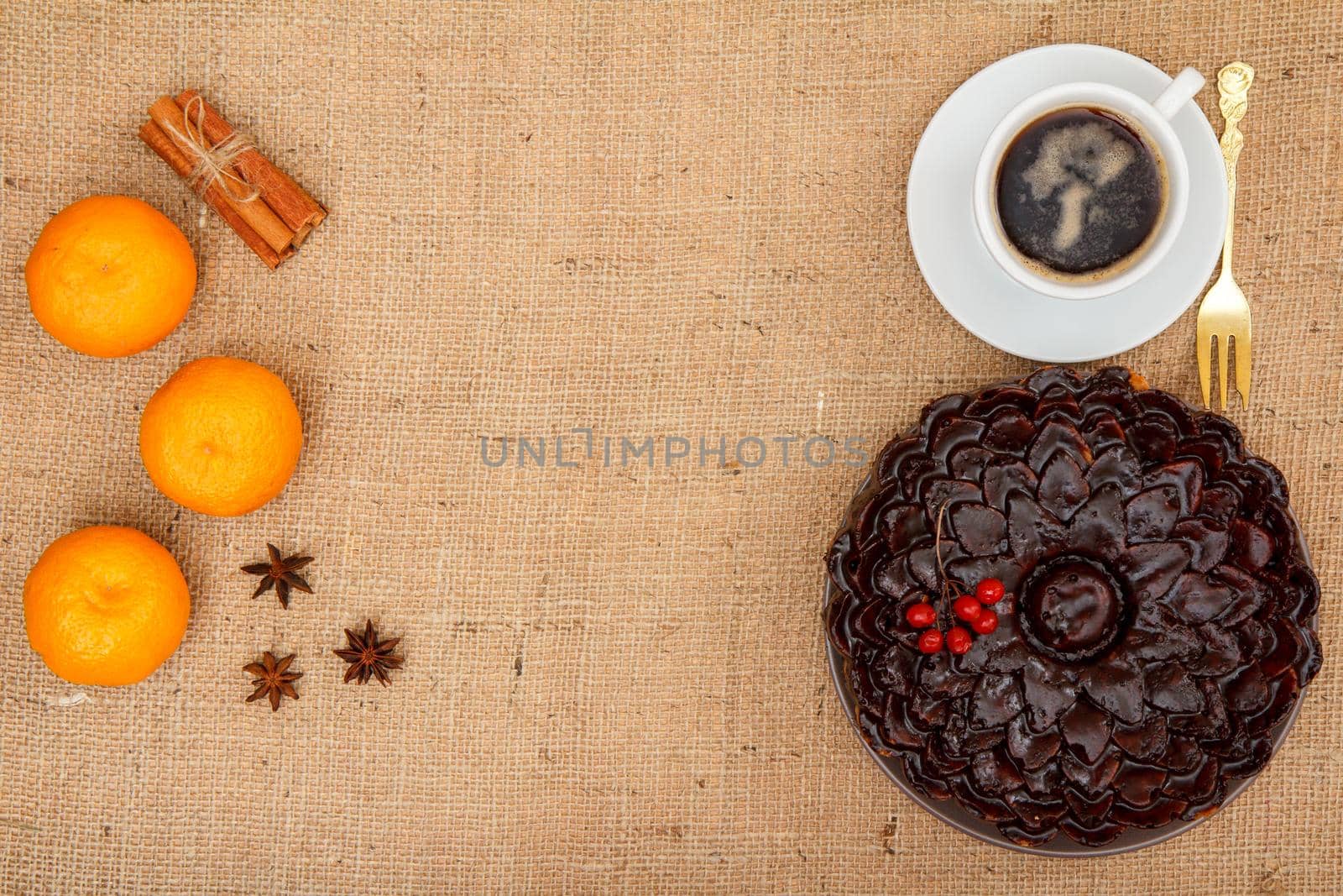 Chocolate cake decorated with bunch of viburnum, fork and cup of coffee beside it, oranges, star anise and cinnamon on table with sackcloth. Top view