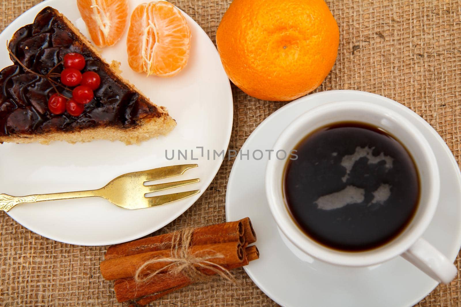 Slice of chocolate cake decorated with bunch of viburnum, fork, cup of coffee, oranges and cinnamon on table with sackcloth. Top view