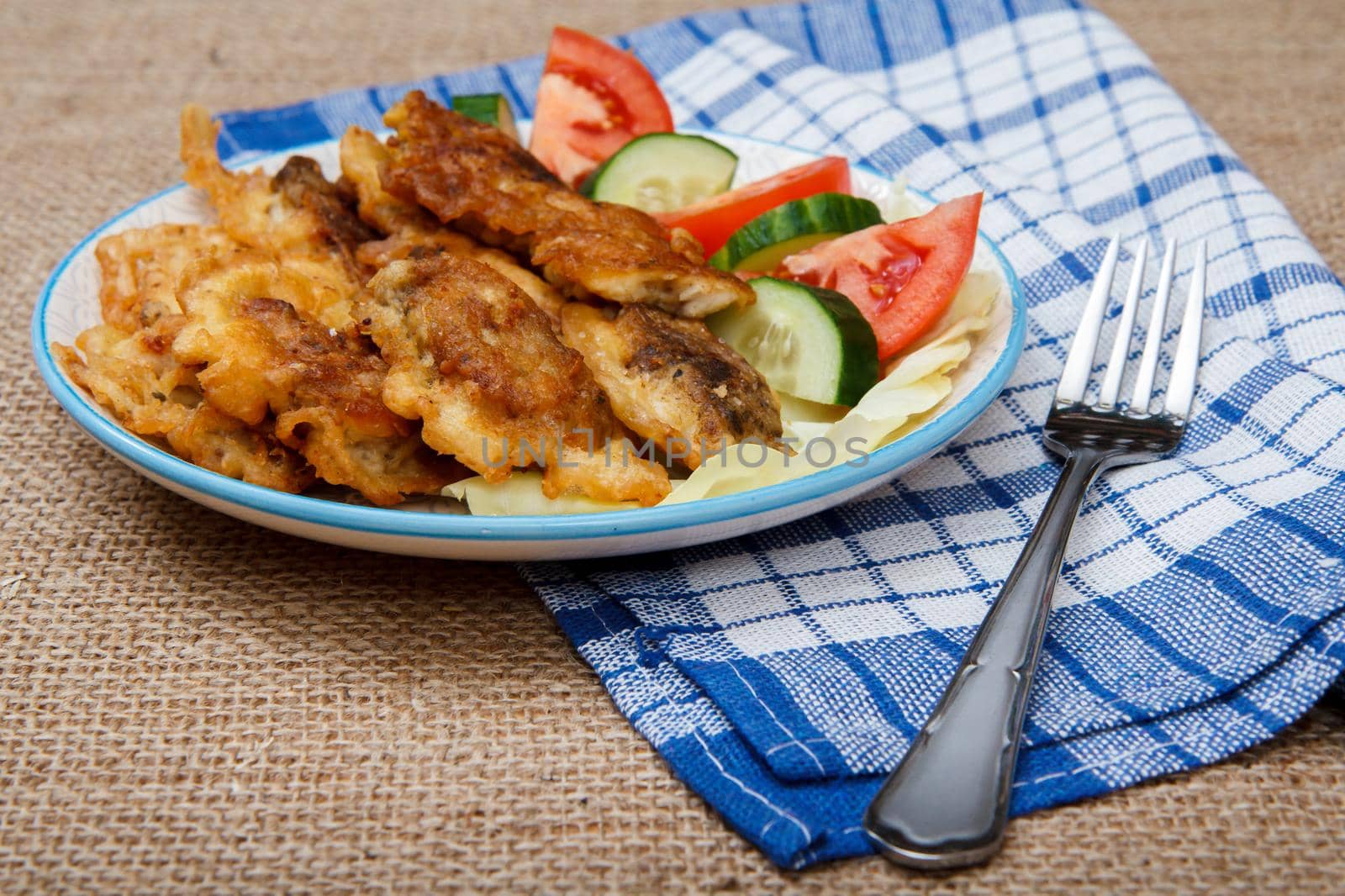 Plate with pieces of fried fish and freshly sliced tomatoes and cucumbers, napkin with fork on sackcloth