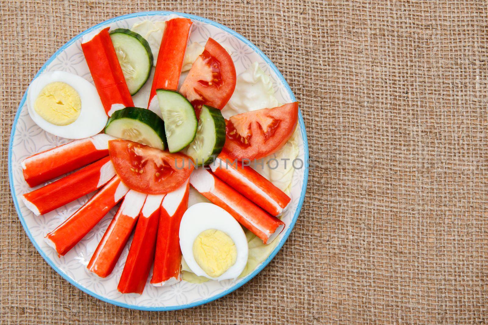 Plate with crab sticks, boiled egg and freshly sliced tomato and cucumber on sackcloth. Top view with copy space