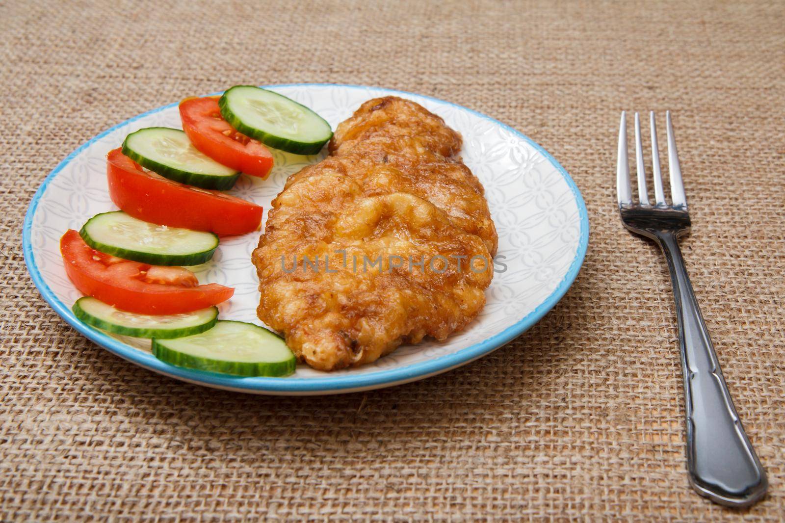 Plate with fried meat chop in batter and sliced tomato and cucumber with fork on sackcloth