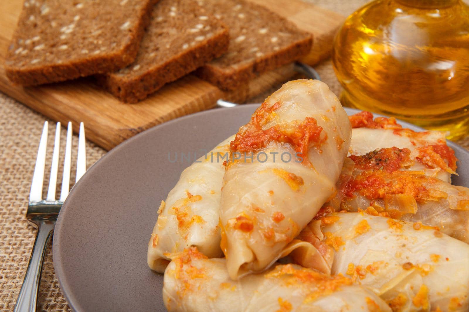 Delicious homemade cabbage rolls stuffed with rice and meat in plate, fork, bottle with sunflower oil and bread on wooden cutting board on sackcloth
