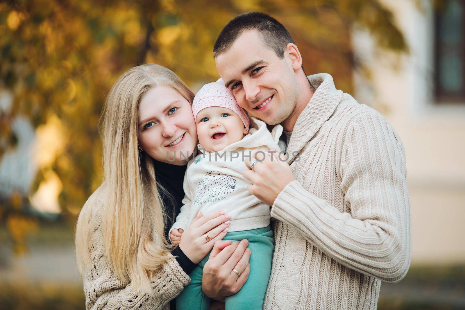 Portrait of attractive husband and blonde wife holding their surprised child. Handsome man helping pretty woman caring about little baby. Young happy family smiling at camera and standing together.