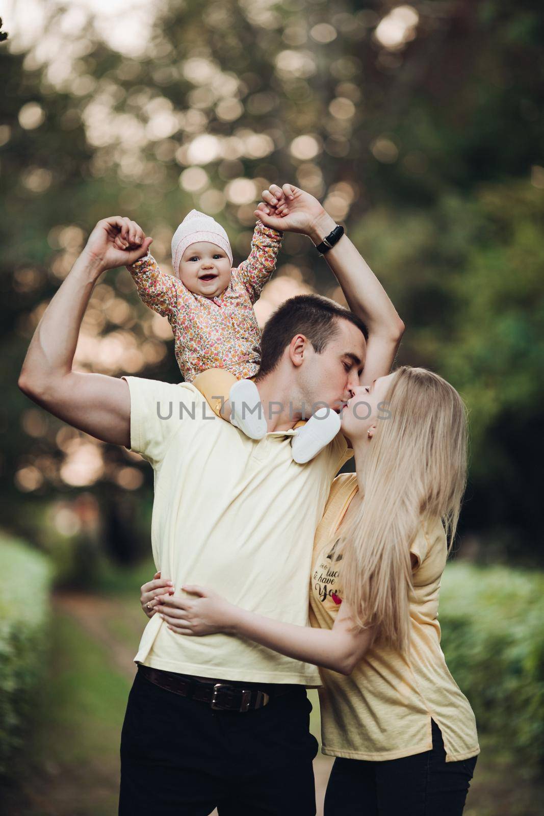 Portrait of lovely young family sitting together outside. Attractive blonde mother smiling with closed eyes. Handsome husband kissing his wife in head and holding happy baby in bright clothes.