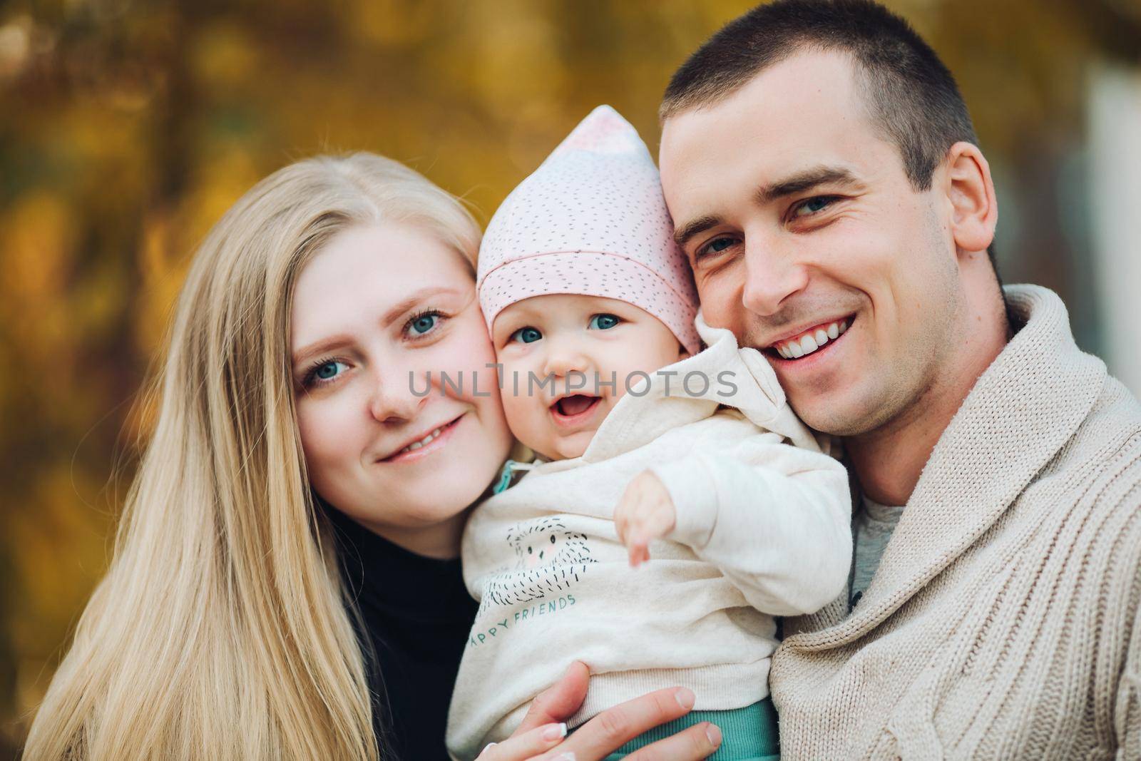 Portrait of attractive husband and blonde wife holding their surprised child. Handsome man helping pretty woman caring about little baby. Young happy family smiling at camera and standing together.