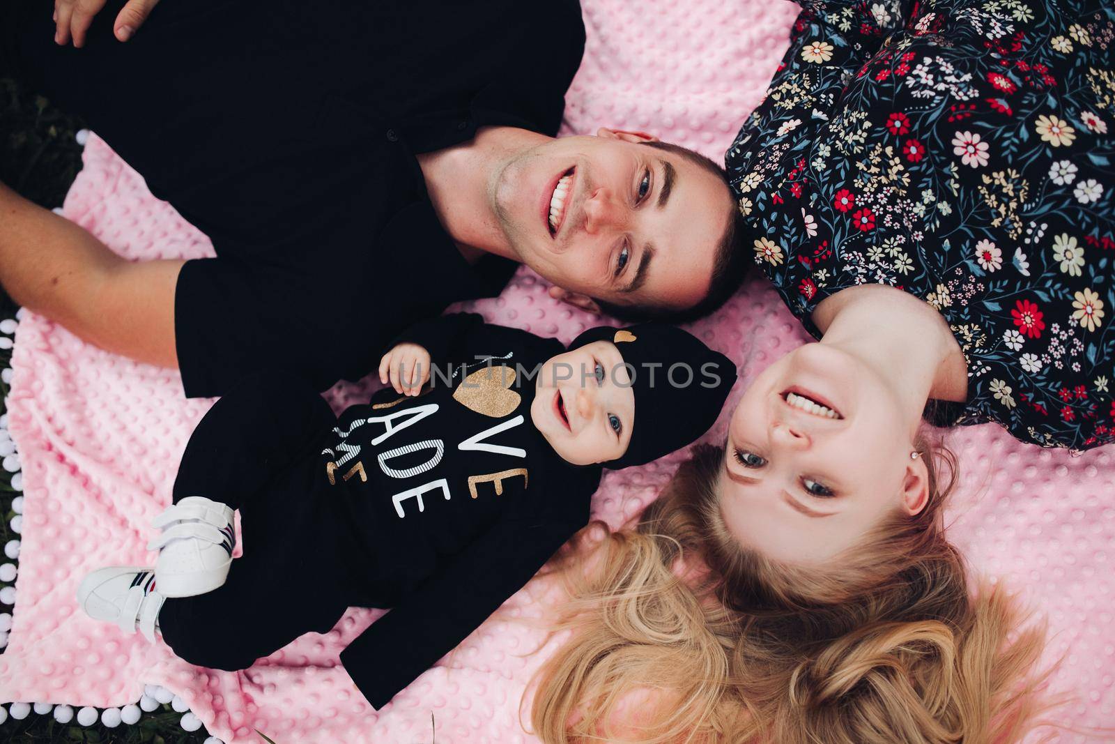 Close up from above of young happy family lying on ground outside. Attractive wife and husband with little baby looking at camera and smiling. Beautiful couple enjoying moment with their lovely child.