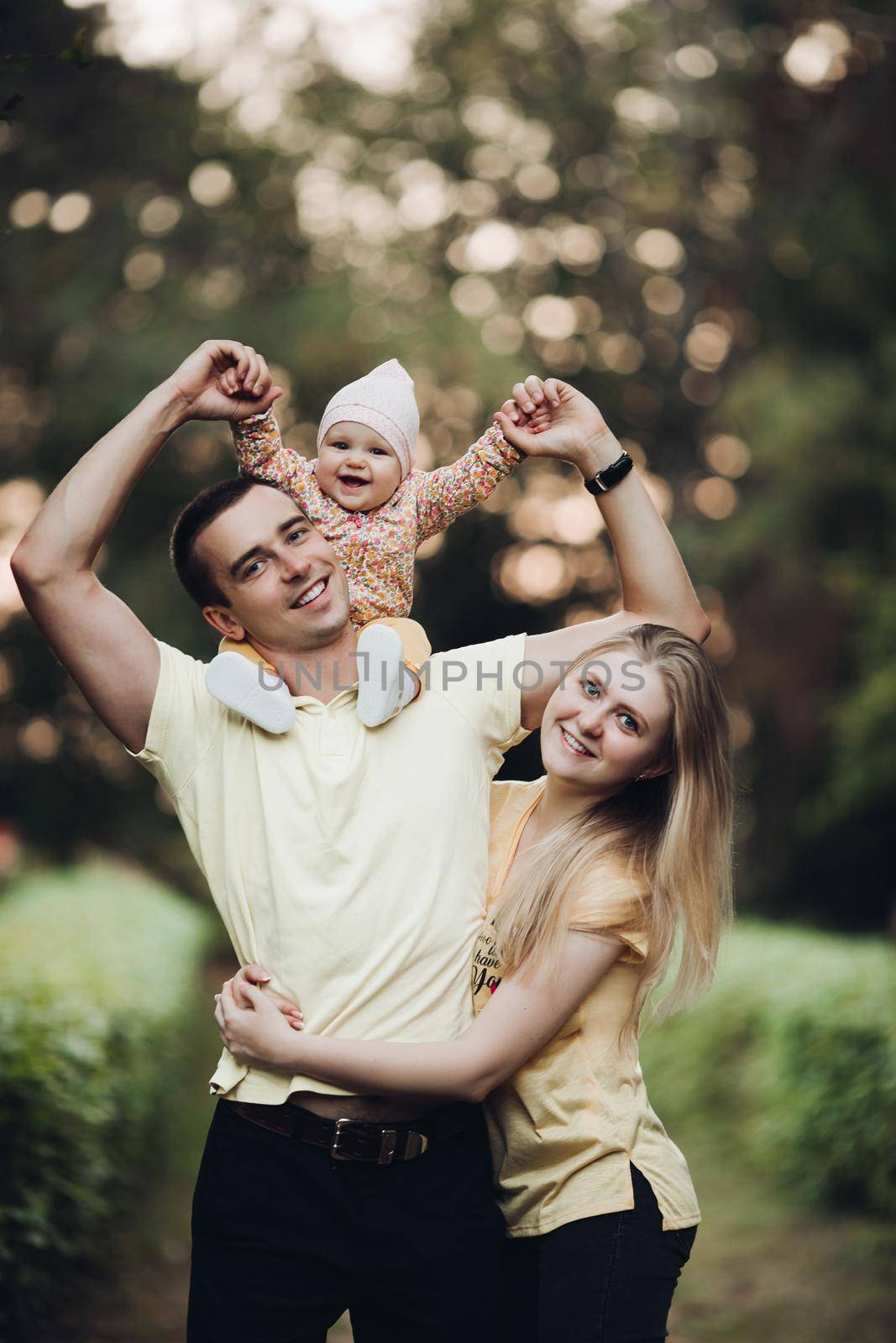 Portrait of lovely young family sitting together outside. Attractive blonde mother smiling with closed eyes. Handsome husband kissing his wife in head and holding happy baby in bright clothes.