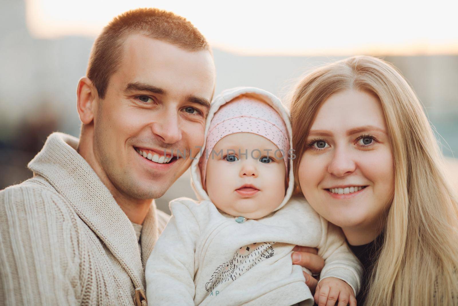 Portrait of attractive husband and blonde wife holding their surprised child. Handsome man helping pretty woman caring about little baby. Young happy family smiling at camera and standing together.