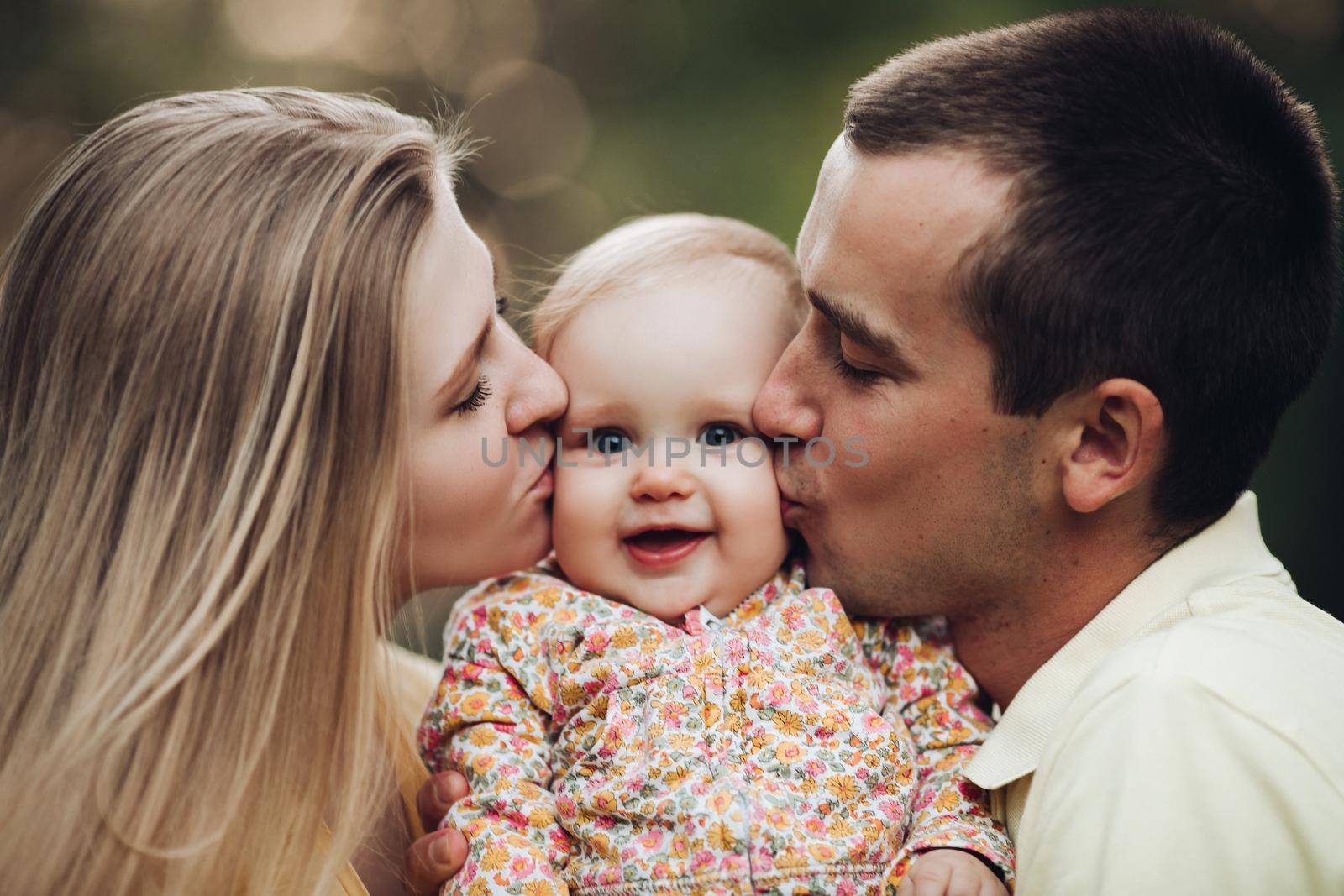 Portrait of lovely young family sitting together outside. Attractive blonde mother smiling with closed eyes. Handsome husband kissing his wife in head and holding happy baby in bright clothes.