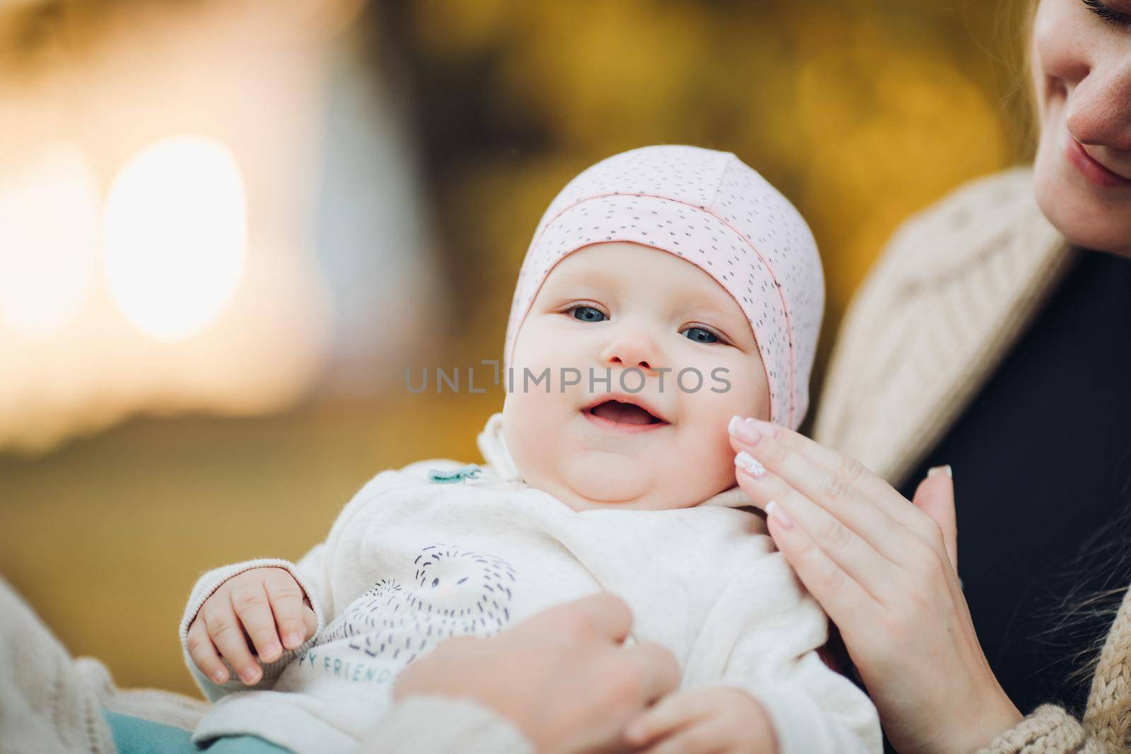 Close up of surprised baby's face with lightly opened mouth. Sweet lovely child in colorful clothes and hat leaning on mother's back. Father's hand holding infant while it looking at camera.