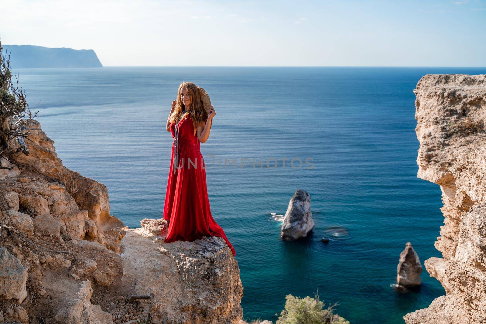 A woman in a flying red dress fluttering in the wind and a straw hat against the backdrop of the sea