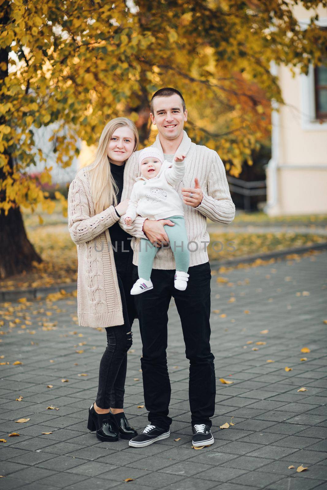 Portrait of lovely young family sitting together outside. Attractive blonde mother smiling with closed eyes. Handsome husband kissing his wife in head and holding happy baby in bright clothes.