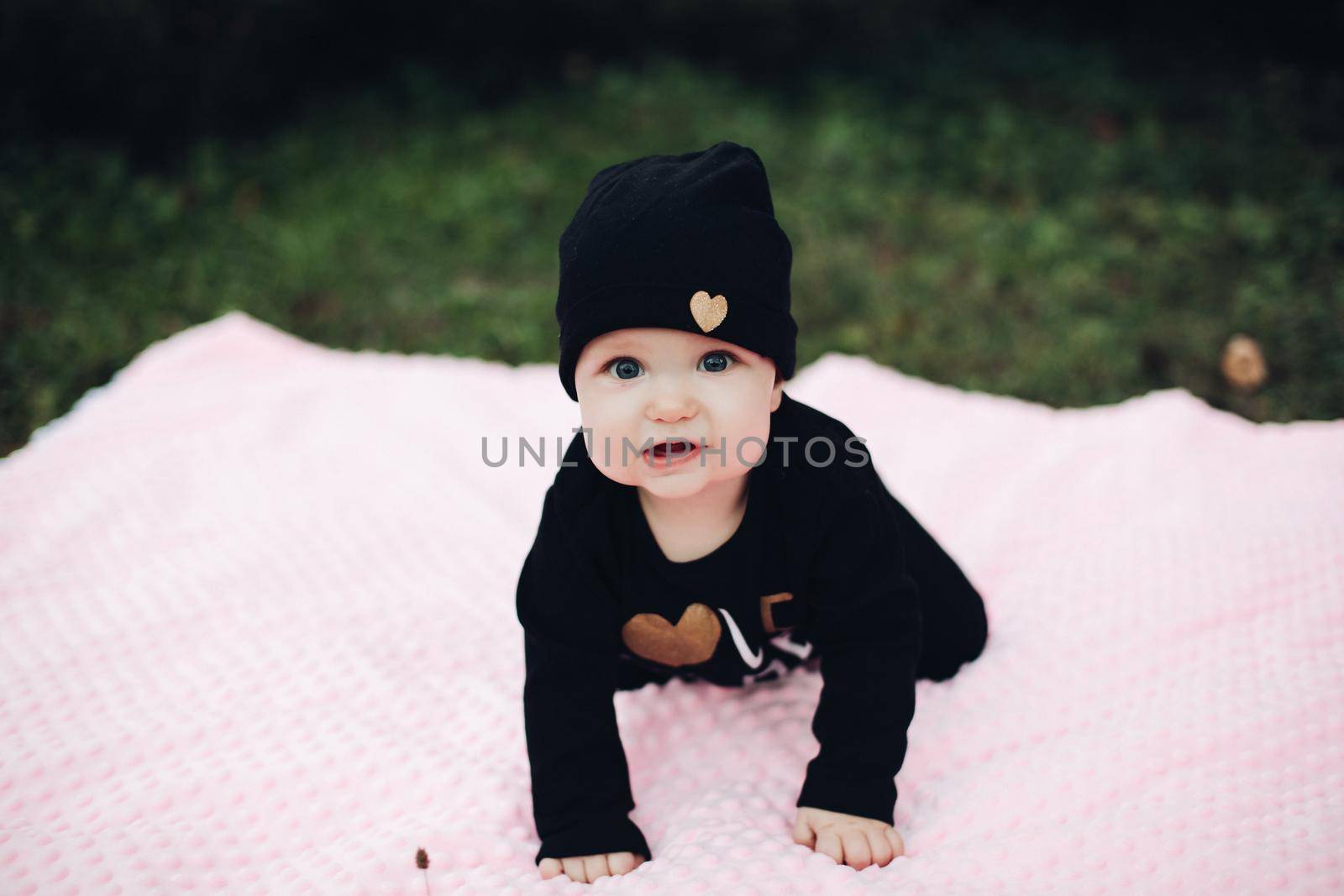 Little kid with cute big eyes lying on pink blanket on ground. Sweet and lovely child with big eyes looking at camera. Stylish infant in total black clothes and hat with gold heart leaning on hands.