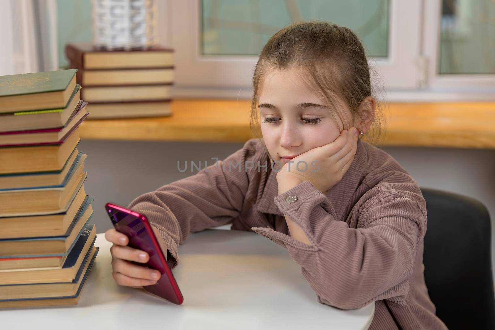 girl sits at a table on which there are books and looks at the phone