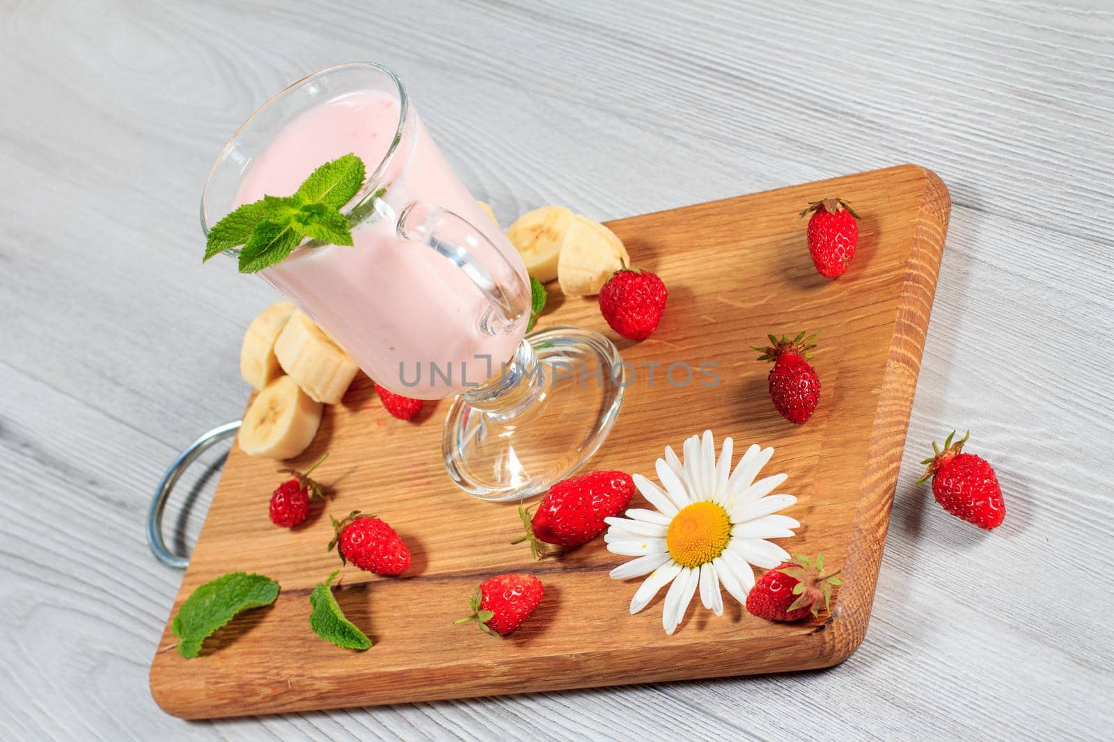 Glass of delicious milk shake with mint and fresh strawberries, banana, chamomile on a wood cutting board