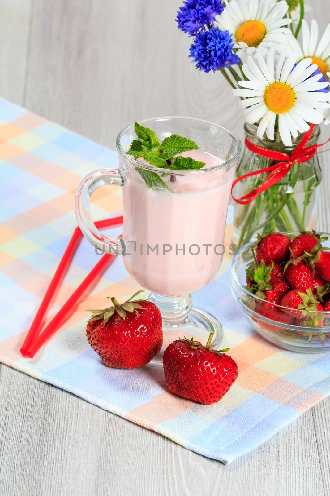 Glass of delicious yogurt with mint and fresh strawberries, chamomile and cornflowers in vase on a wooden table with a napkin