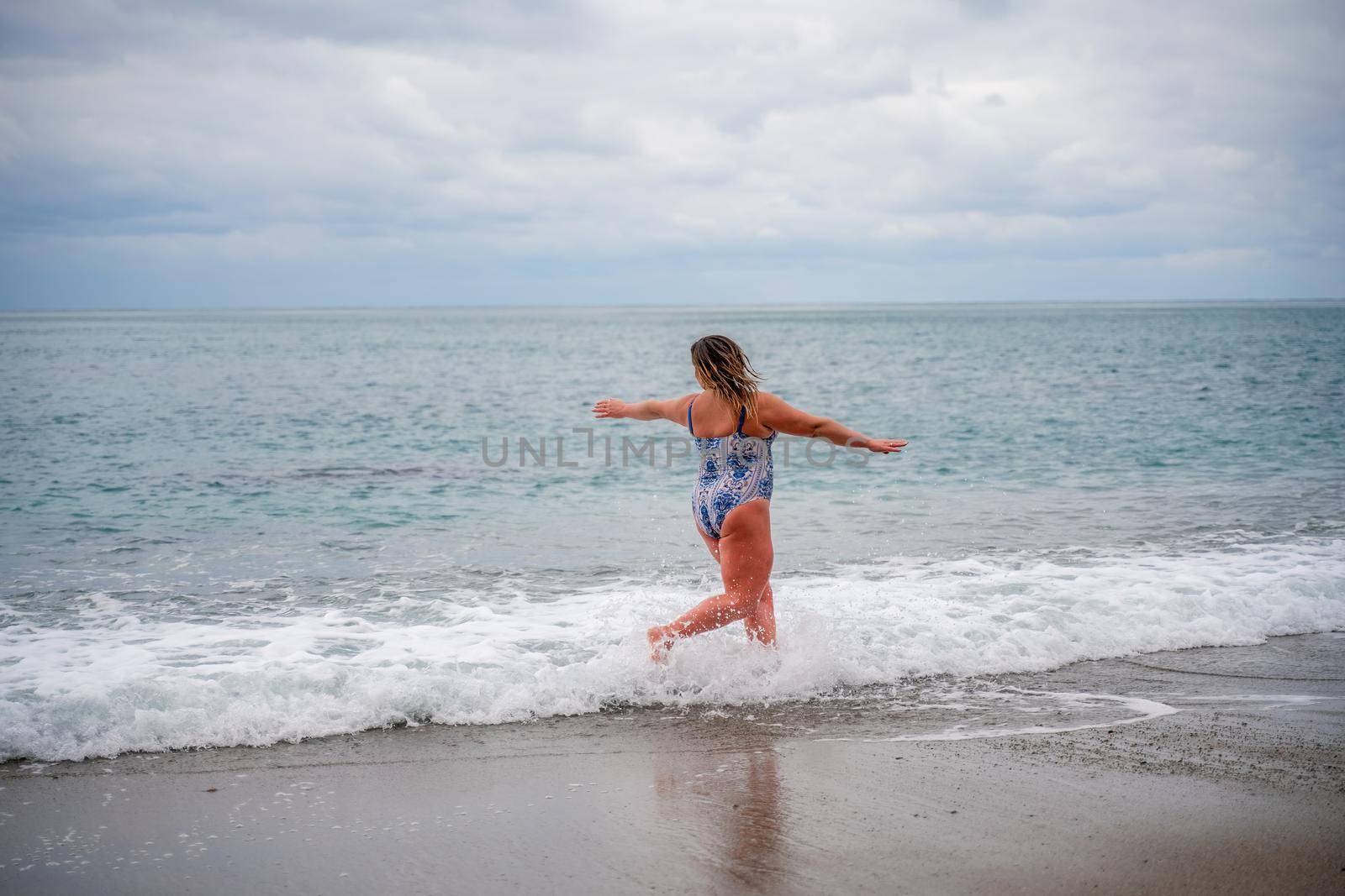 A plump woman in a bathing suit enters the water during the surf. Alone on the beach, Gray sky in the clouds, swimming in winter