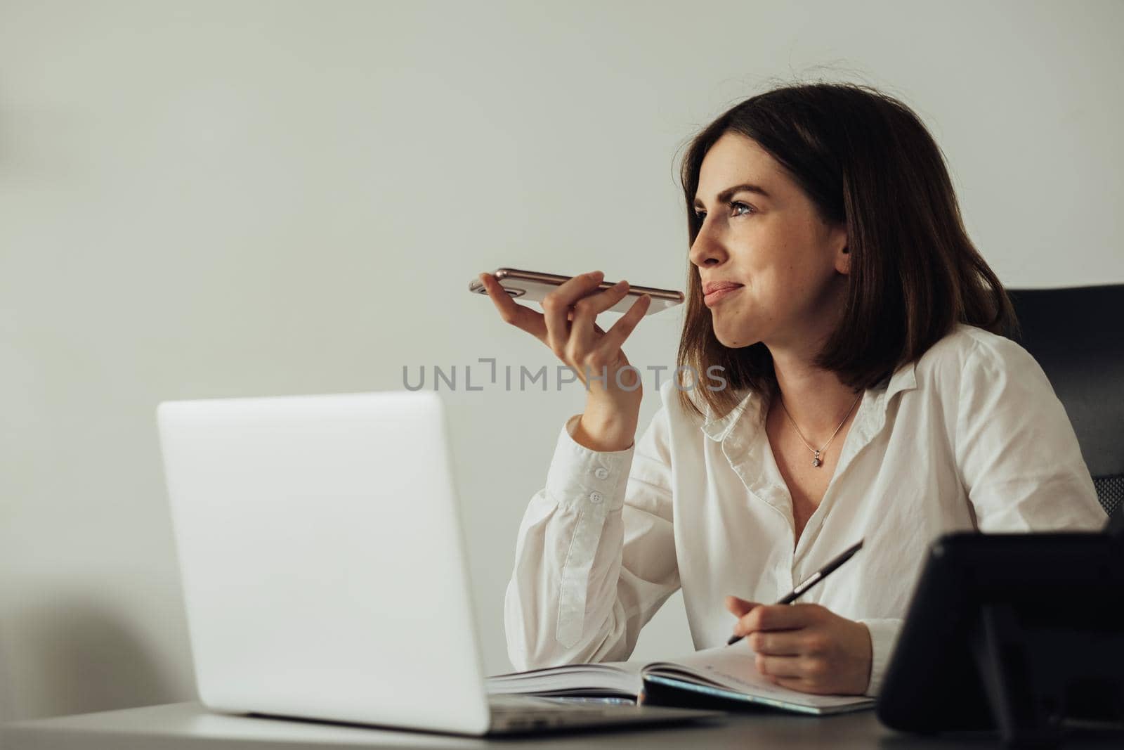 Cheerful Young Woman Working at Office, Female Employee Using Smartphone Voice Message While Sitting by Laptop at Workplace