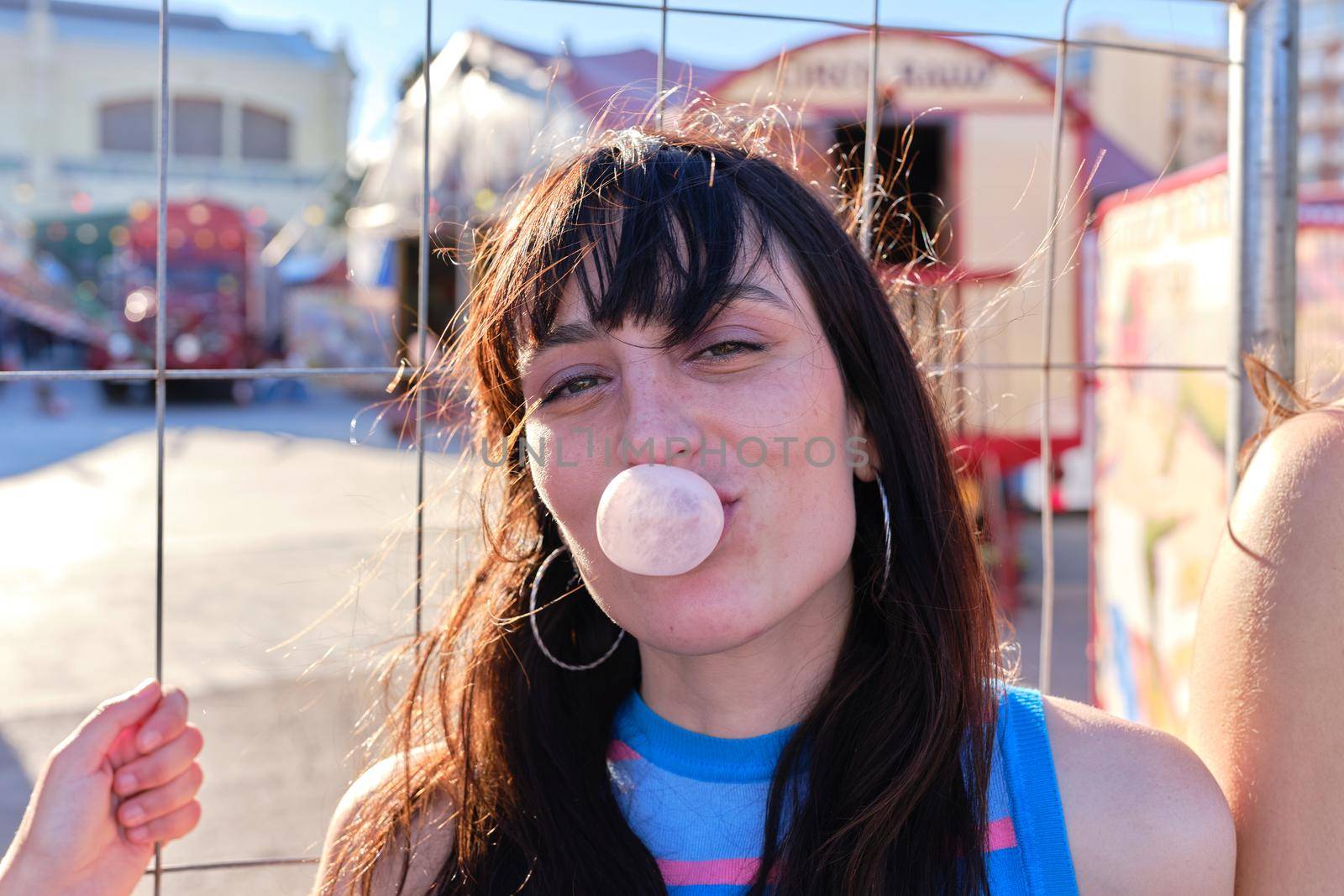 Young beauty woman blowing a bubble with chewing gum next to a circus parade in summer