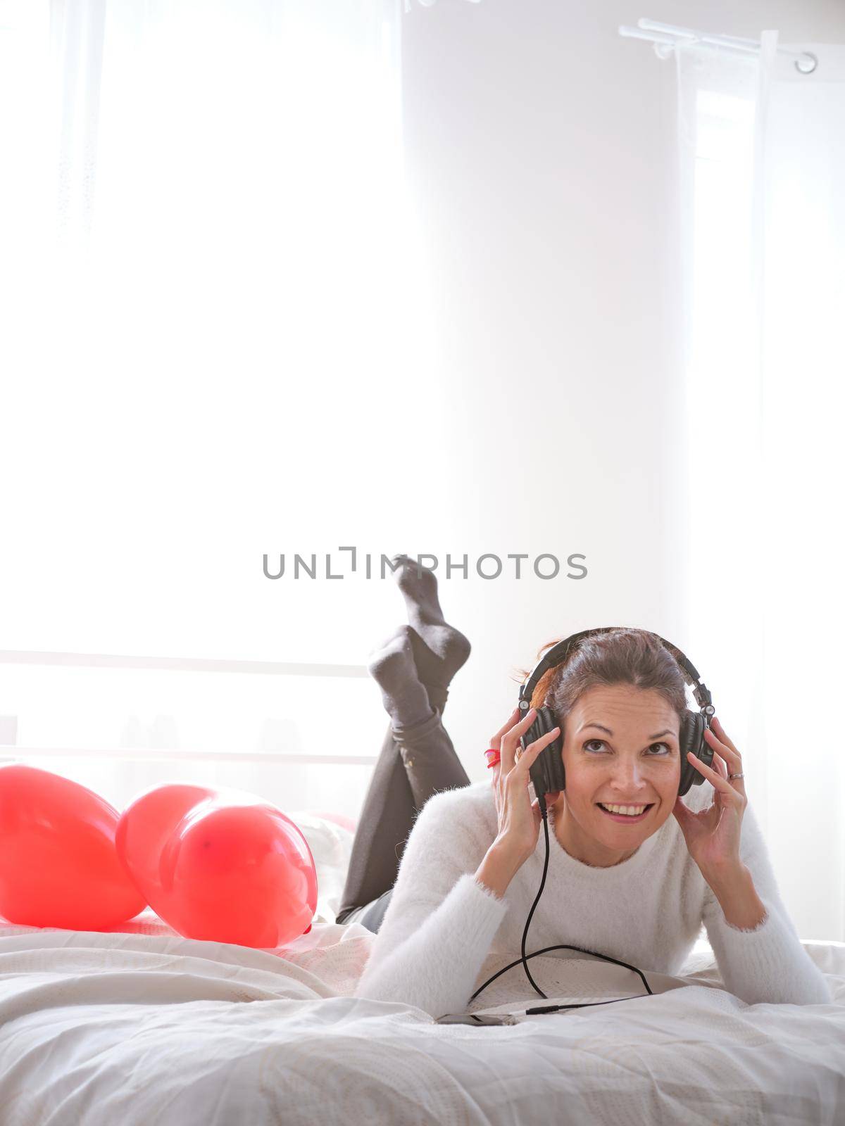 Vertical photo of smiling caucasian woman lying on the bed while listening to music with mobile