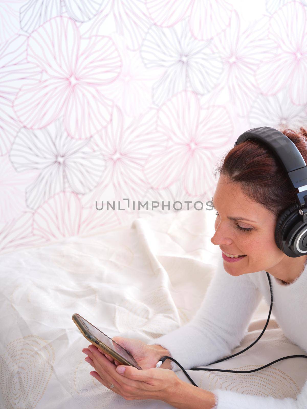 Top view vertical photo of a smiling woman using the mobile while listening to music lying in bed