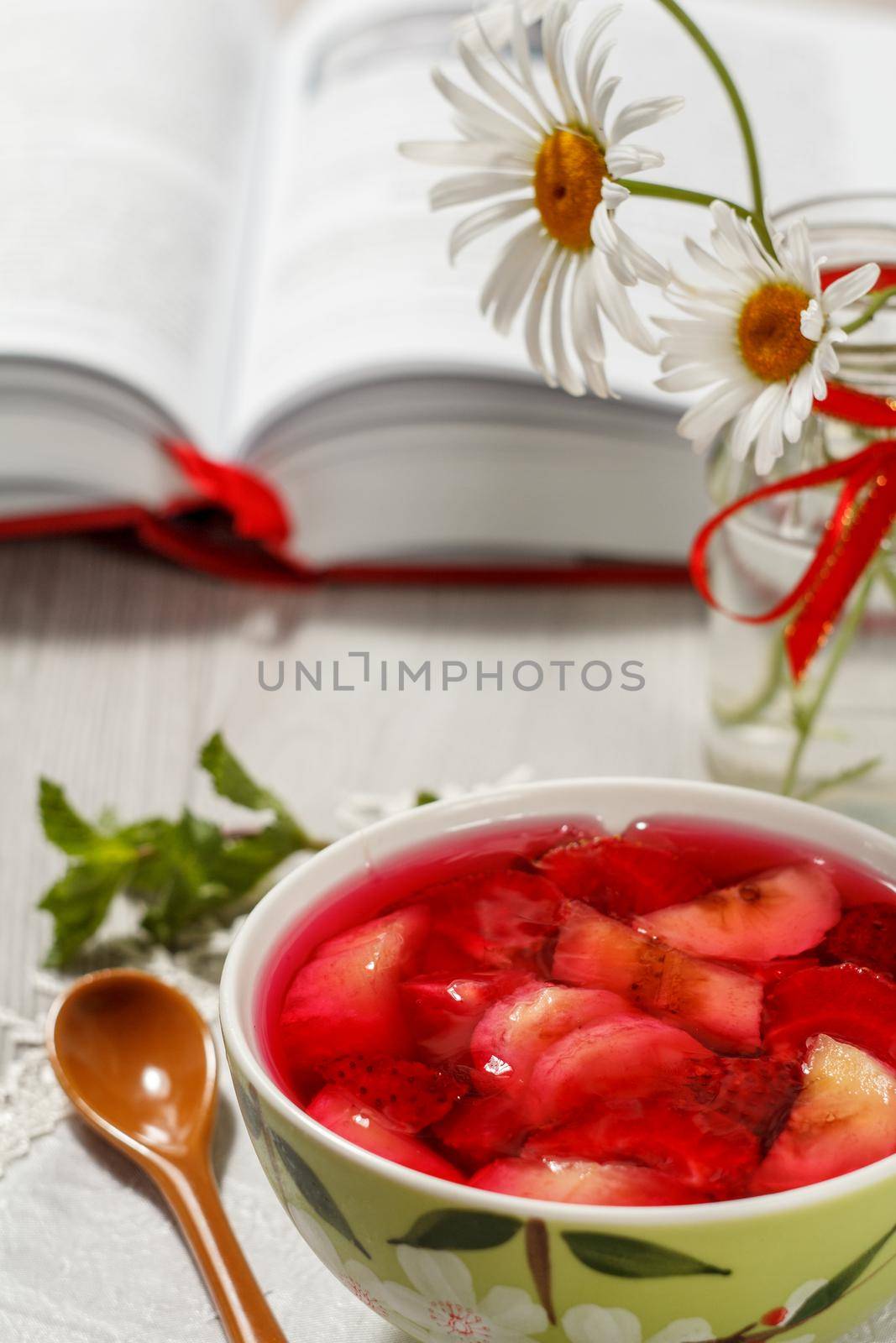 Cherry jelly with strawberry pieces in the bowl with opened book and chamomiles on the background