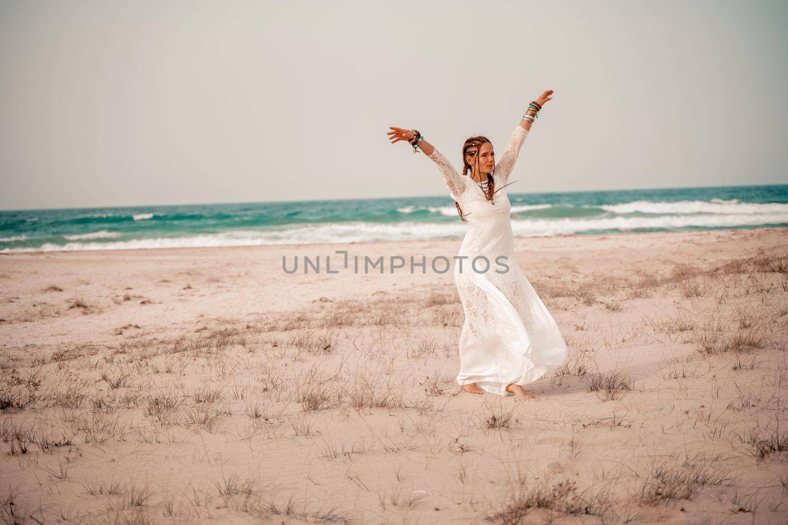 Model in boho style in a white long dress and silver jewelry on the beach. Her hair is braided, and there are many bracelets on her arms. by Matiunina