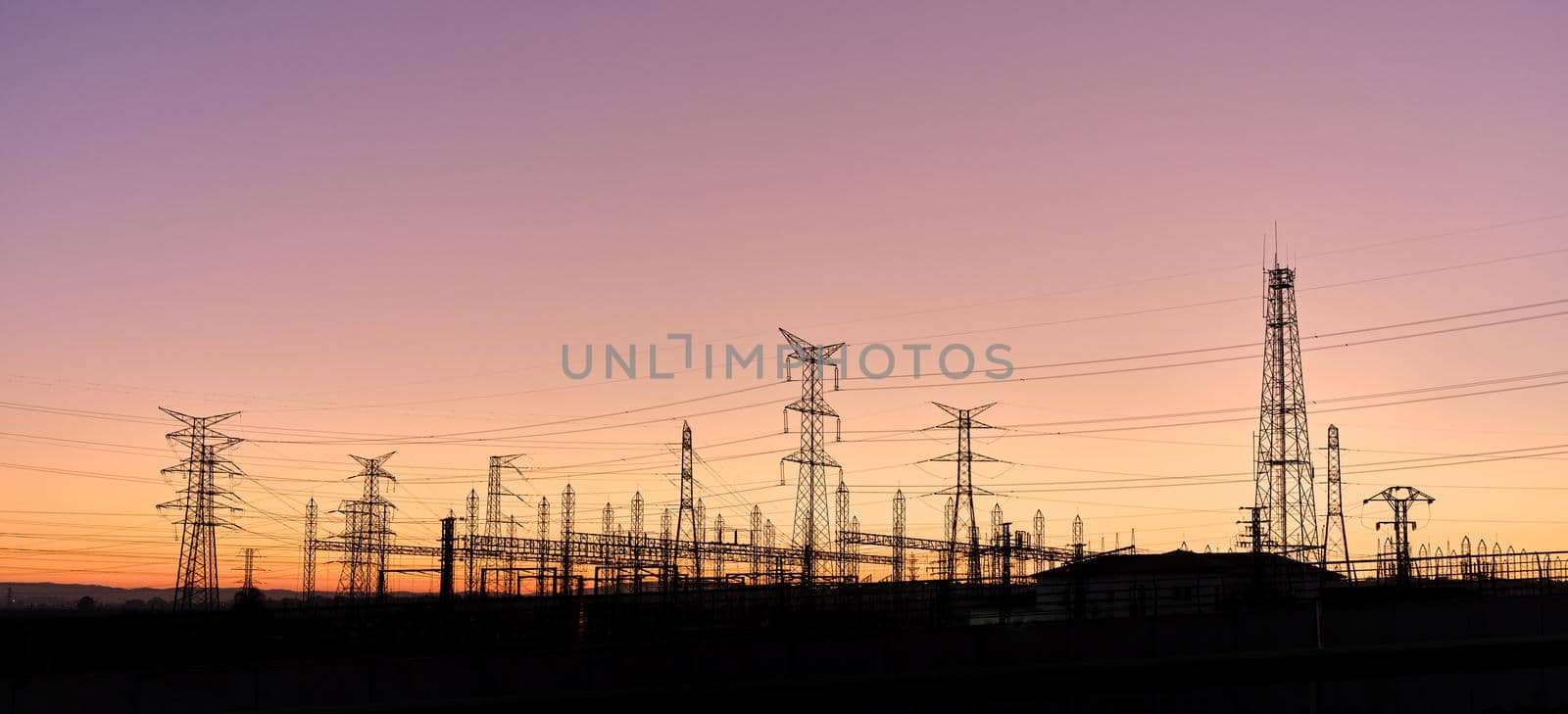 Panoramic photo of silhouettes of several electric towers connected by electric wires during sunset