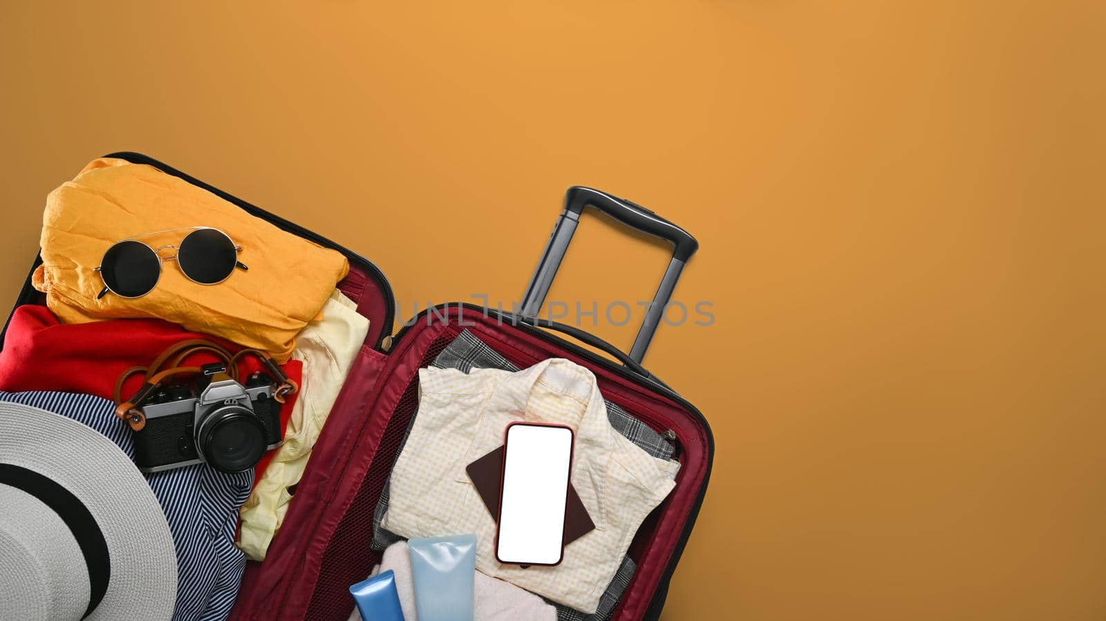 Open suitcase with smart phone, sunglasses, camera and clothes on yellow background. Preparing for the summer vacations.