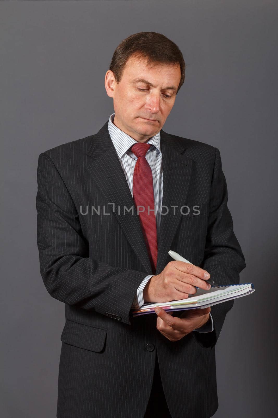 Confident and friendly elegant handsome mature businessman standing in front of a gray background holding a marker and writing in a notebook