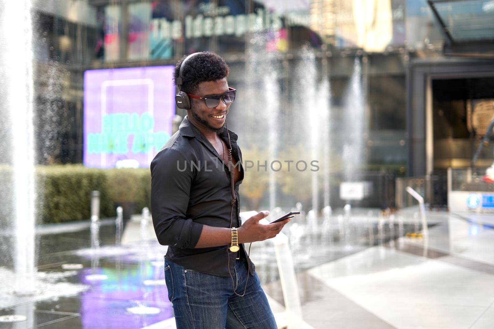 Stylish african man smiling while listening to music with a mobile outside a mall in Bangkok