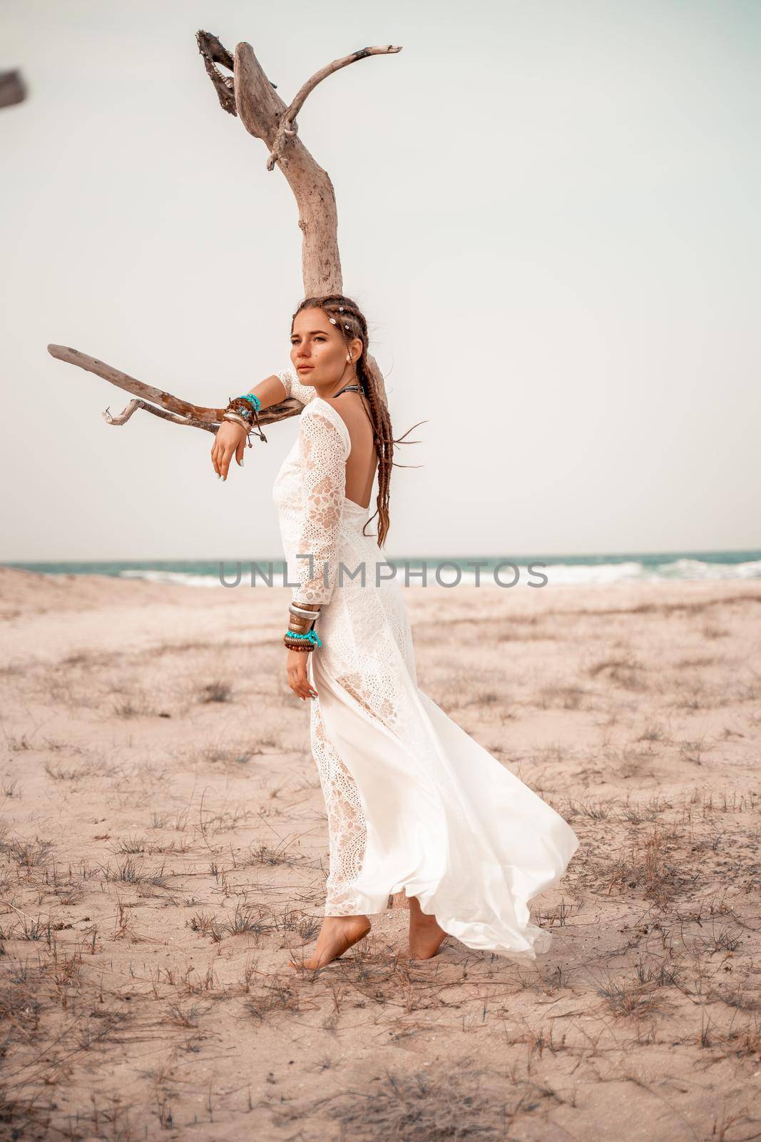 Model in boho style in a white long dress and silver jewelry on the beach. Her hair is braided, and there are many bracelets on her arms