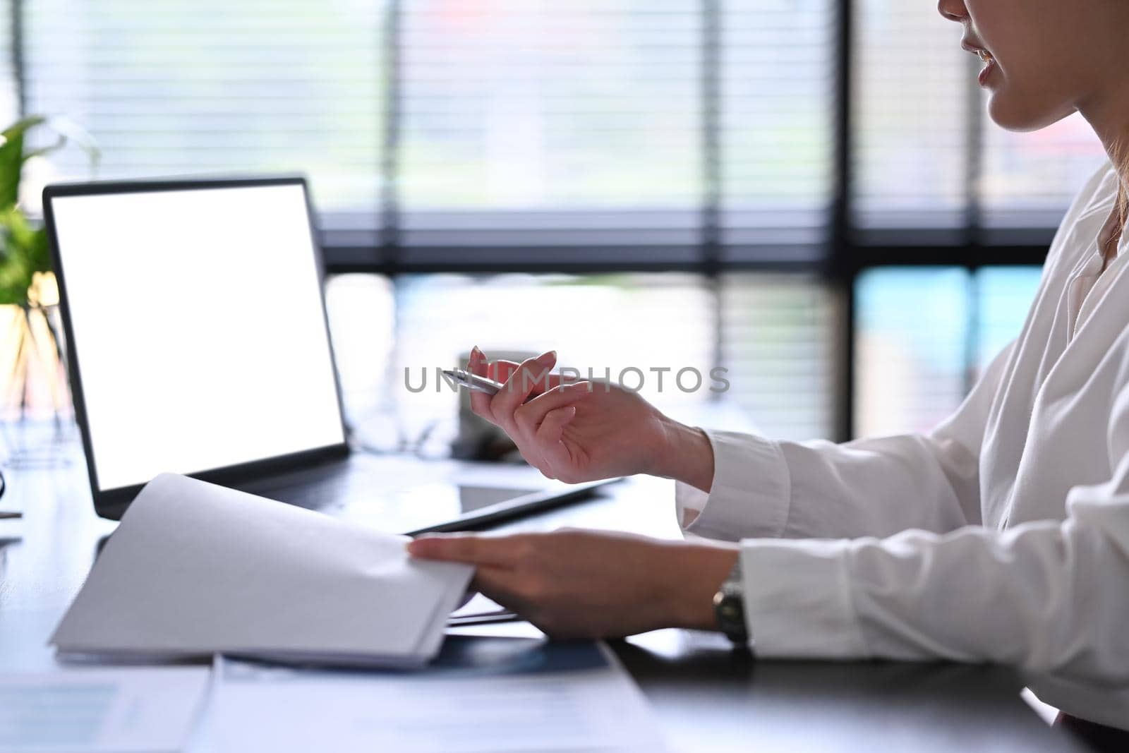 Cropped shot female office worker holding document and using laptop computer at modern workplace.
