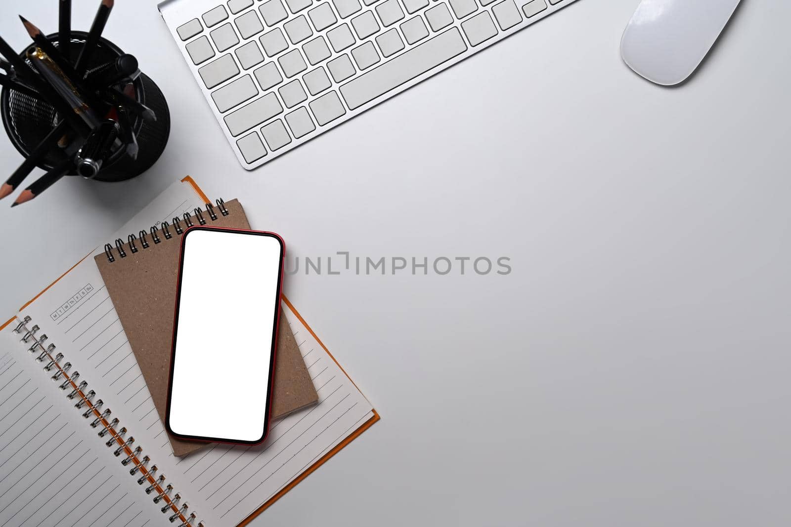 Top view smart phone, notebook, pencil holder and wireless keyboard on white table.