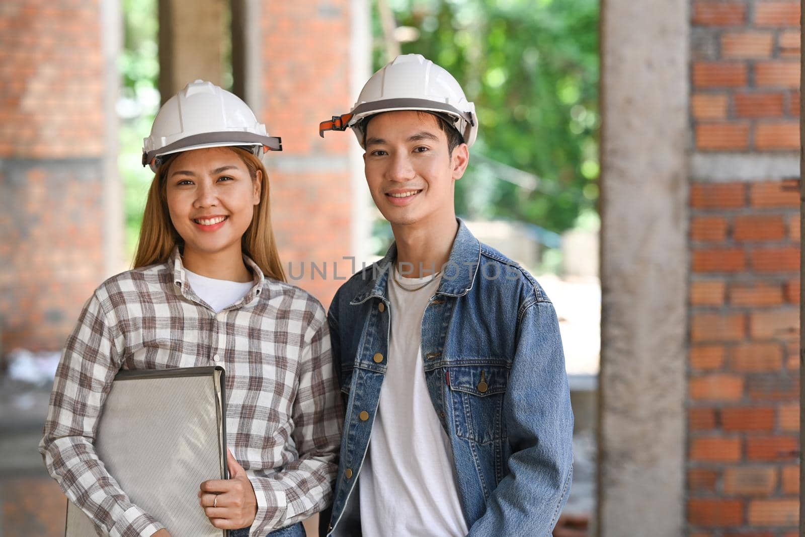 Two civil engineers wearing safety hard hat standing at building construction site and smiling to camera.