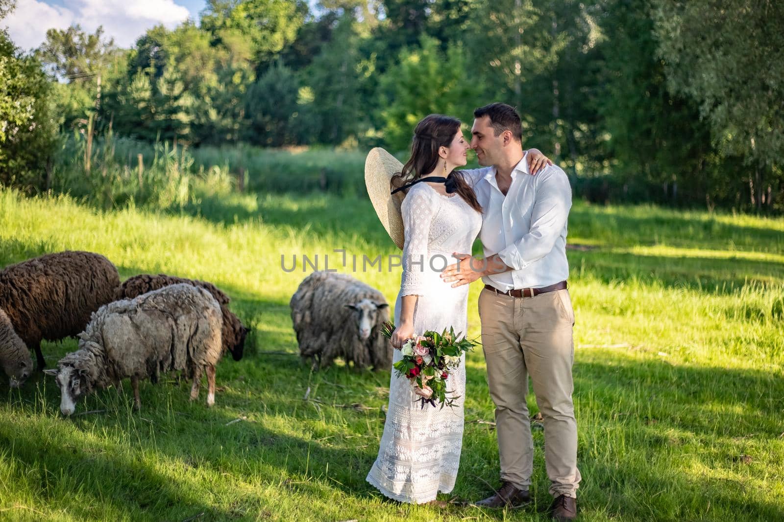 Beautiful newlyweds stand against the background of a fence with green foliage. The stylish bridegroom holds the bride's hand. Wedding