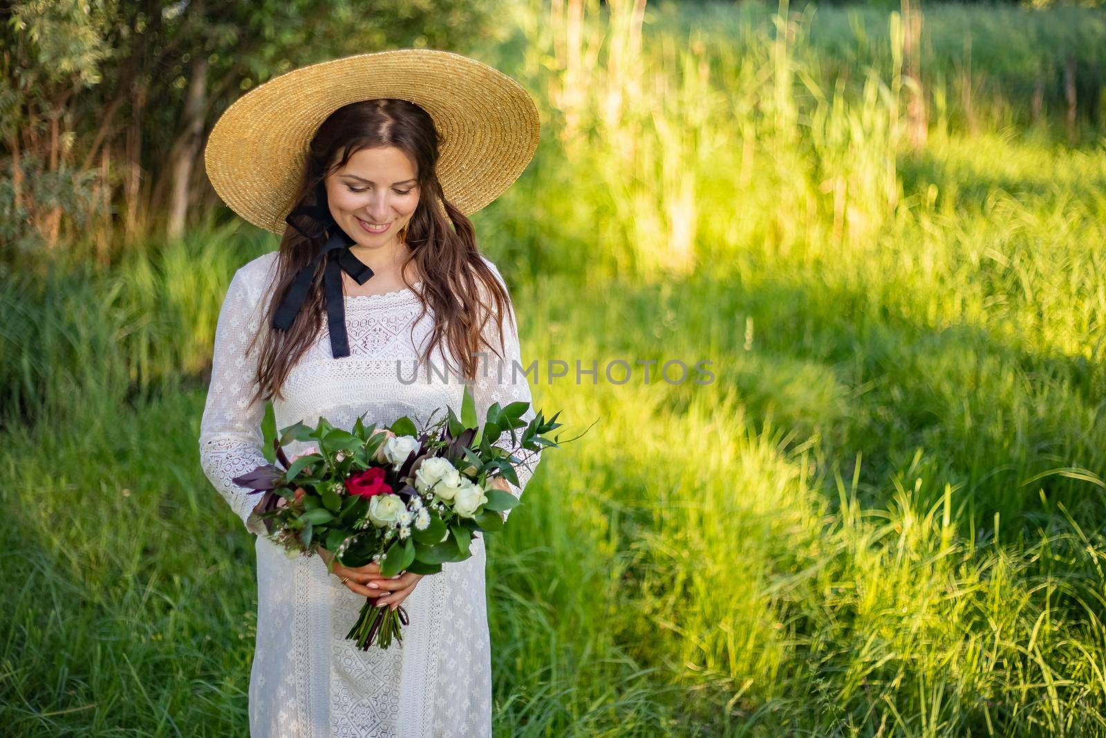 a young, beautiful bride with make-up and hairstyle stands in a meadow in a boho style wedding dress. holding a bouquet of flowers in her hands. large straw hat on shoulder. by Anyatachka