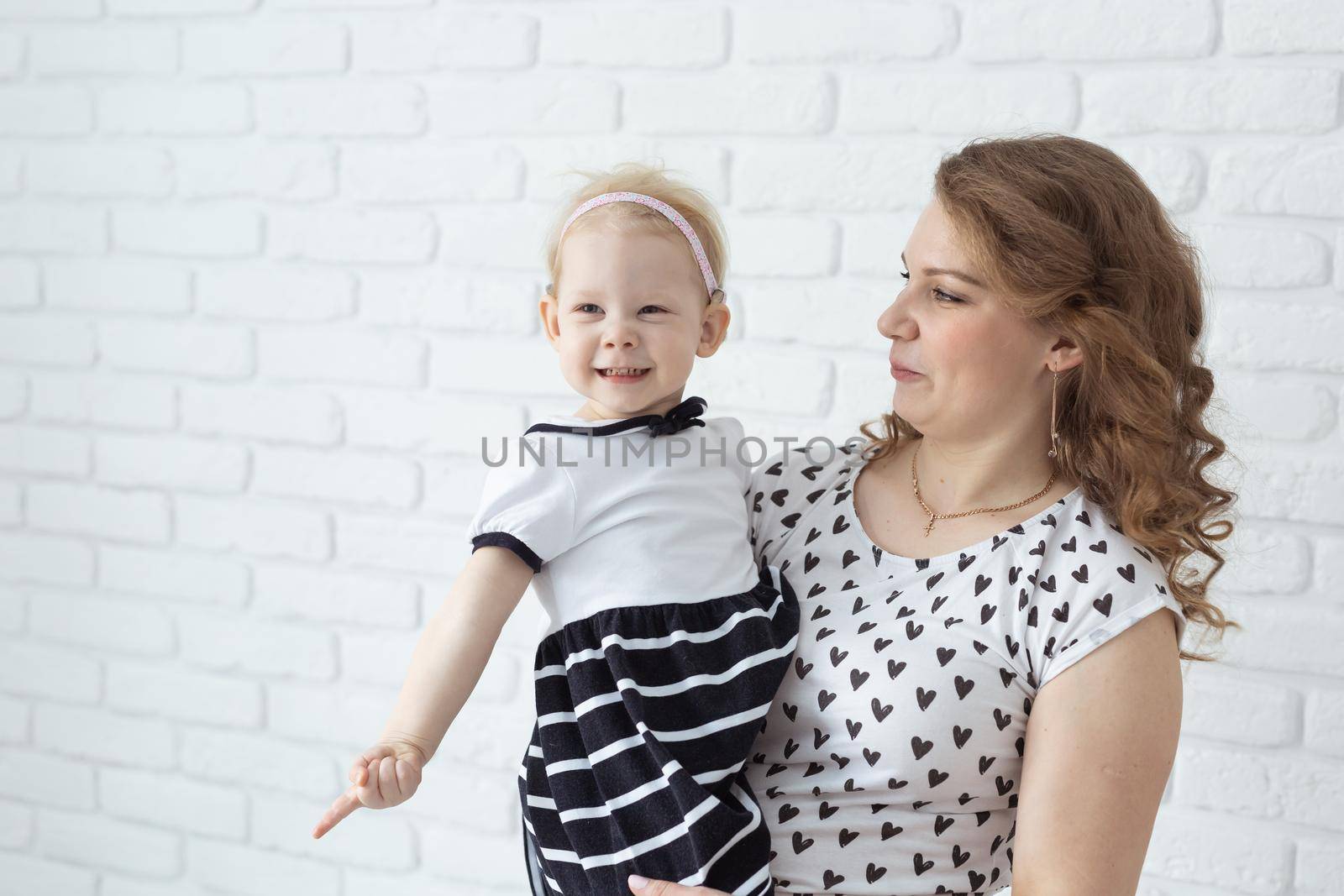 Mother holds and having fun her child with hearing aids and cochlear implants on white brick wall with copy space and place for advertising . Deaf and innovating medicine technologies concept by Satura86