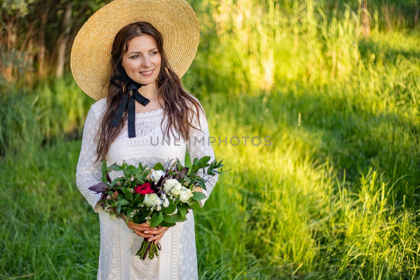 a young, beautiful bride with make-up and hairstyle stands in a meadow in a boho style wedding dress. holding a bouquet of flowers in her hands. large straw hat on shoulder. by Anyatachka