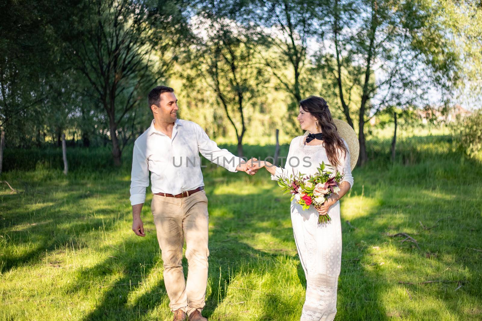 Happy young people in wedding attire walk in the meadow and look at each other by Anyatachka