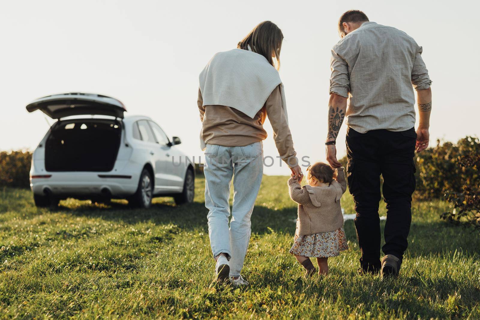 Young Family Enjoying Road Trip on SUV Car, Mother and Tattooed Father Helping Their Baby Daughter Making First Steps Outdoors in Field at Sunset