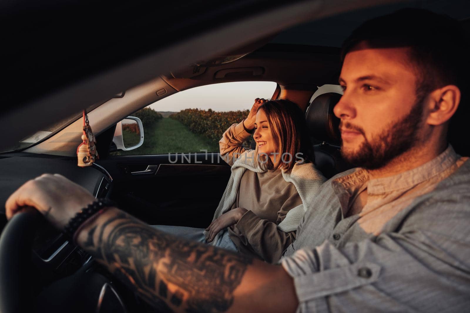 Tattooed Caucasian Man and Young Woman Sitting in Car at Sunset, Couple enjoying Road Trip on Weekend