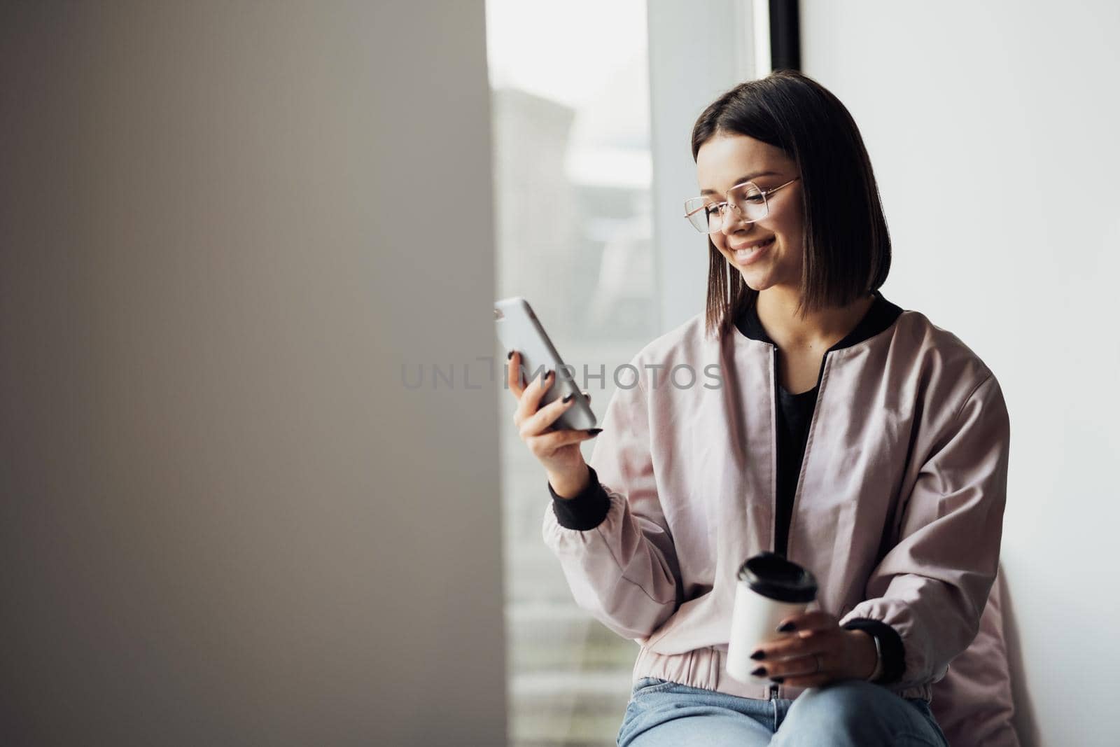Young Woman in Glasses Drinking Coffee and Using Smartphone While Sitting on Windowsill