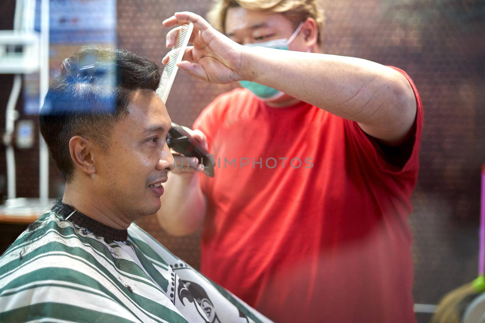 Profile of a fat asian barber with facial mask cutting the hair of an asian client in a barber shop