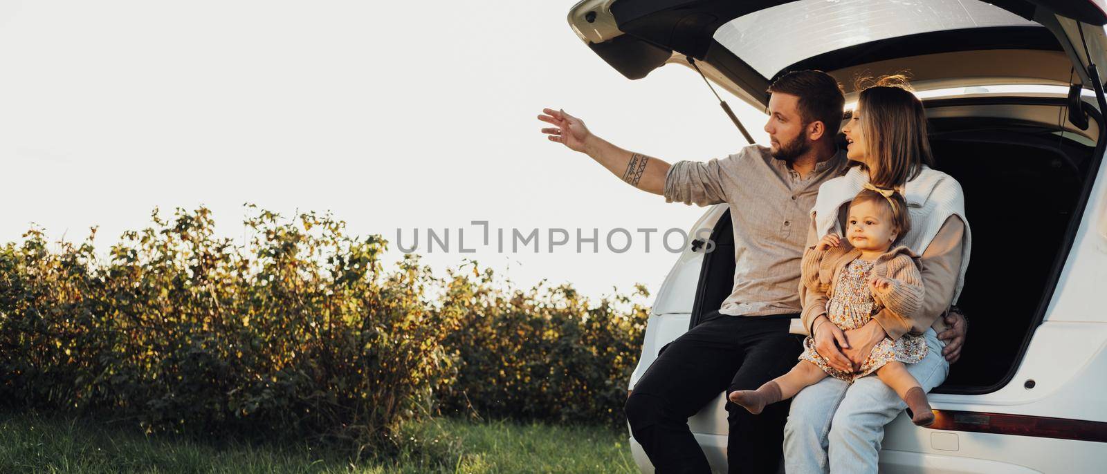 Young Caucasian Family Sitting in Trunk of SUV Car, Mom and Dad with Baby Daughter Having Fun Time on Weekend Outdoors, Copy Space