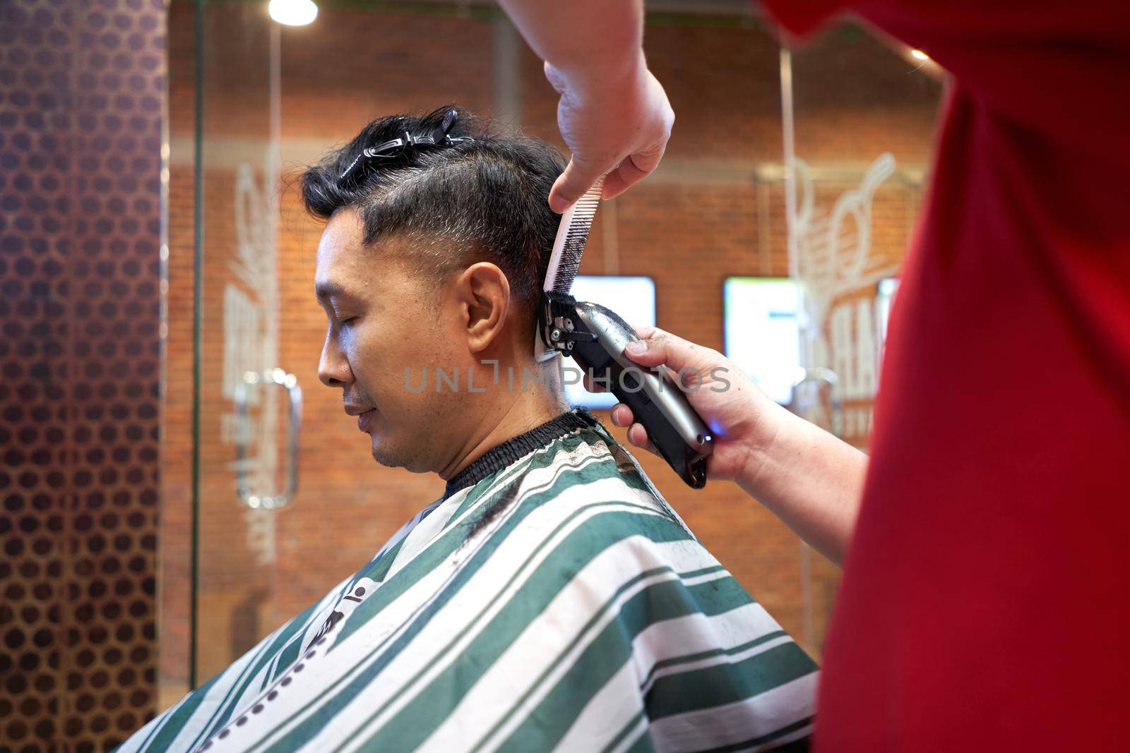 Asian man sits in a barber's shop while his hair is cut with an electric razor. by WesternExoticStockers