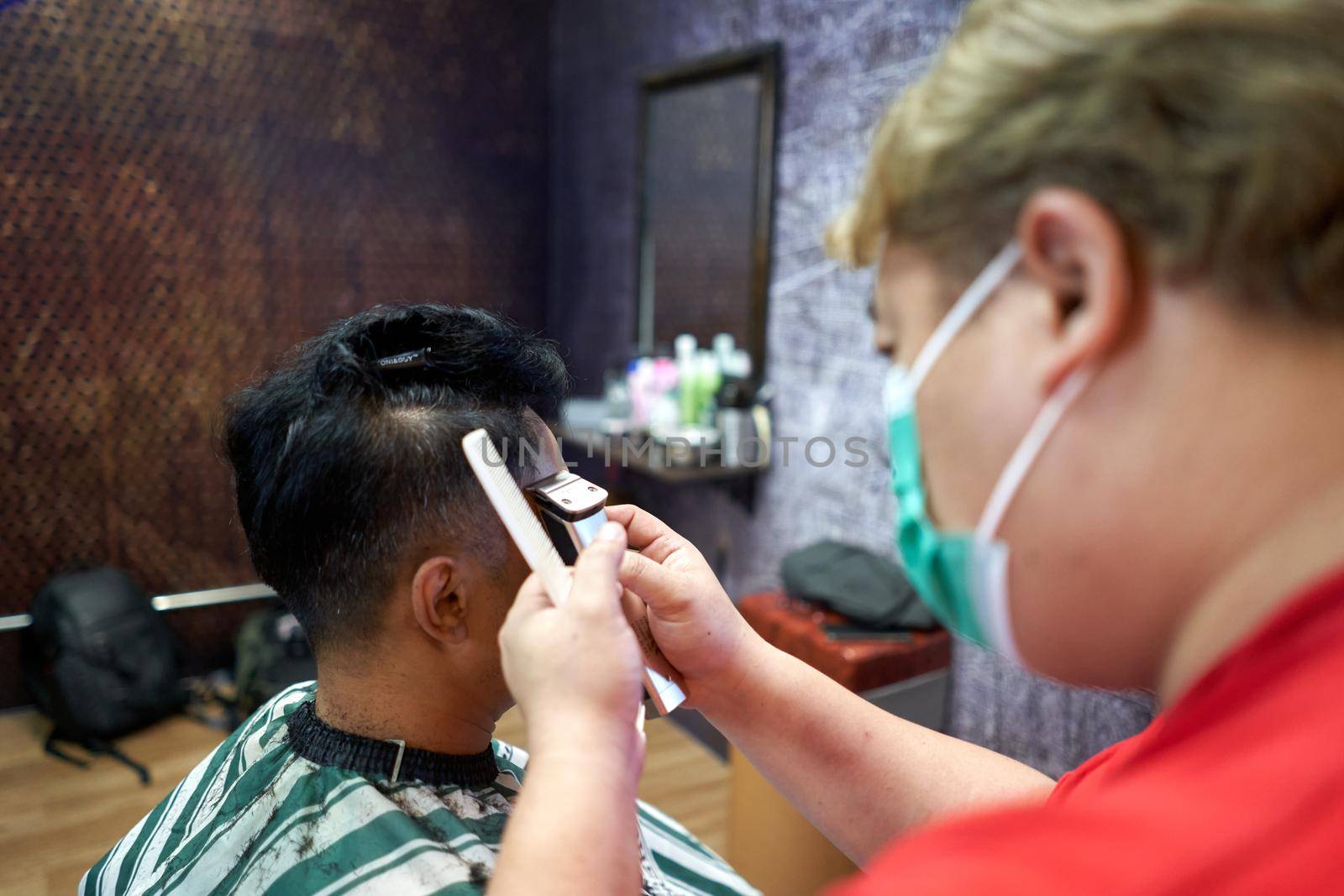Barber with mask cutting the hair on the side of the head of a gay customer sitting in a barbershop