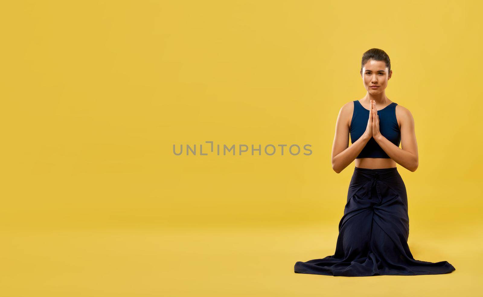 Female yoga instructor sitting on knees in prayer pose in spacious studio. Front view of brunette girl kneeling and praying, looking at camera, isolated on orange studio background. Concept of pray.